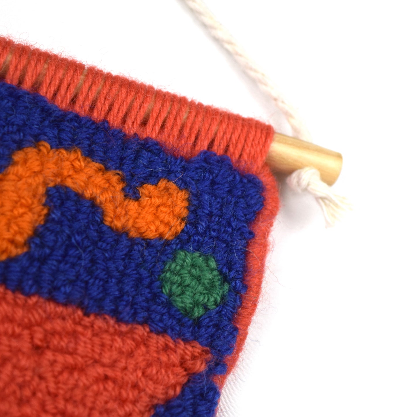 A hand punched miniature, using simple semi circles, circles and squiggle to create a carefully balanced wall hanging using British Wool. Royal Blue, Yellow, Orange, Green and Coral with Coral hand bound edges close up.