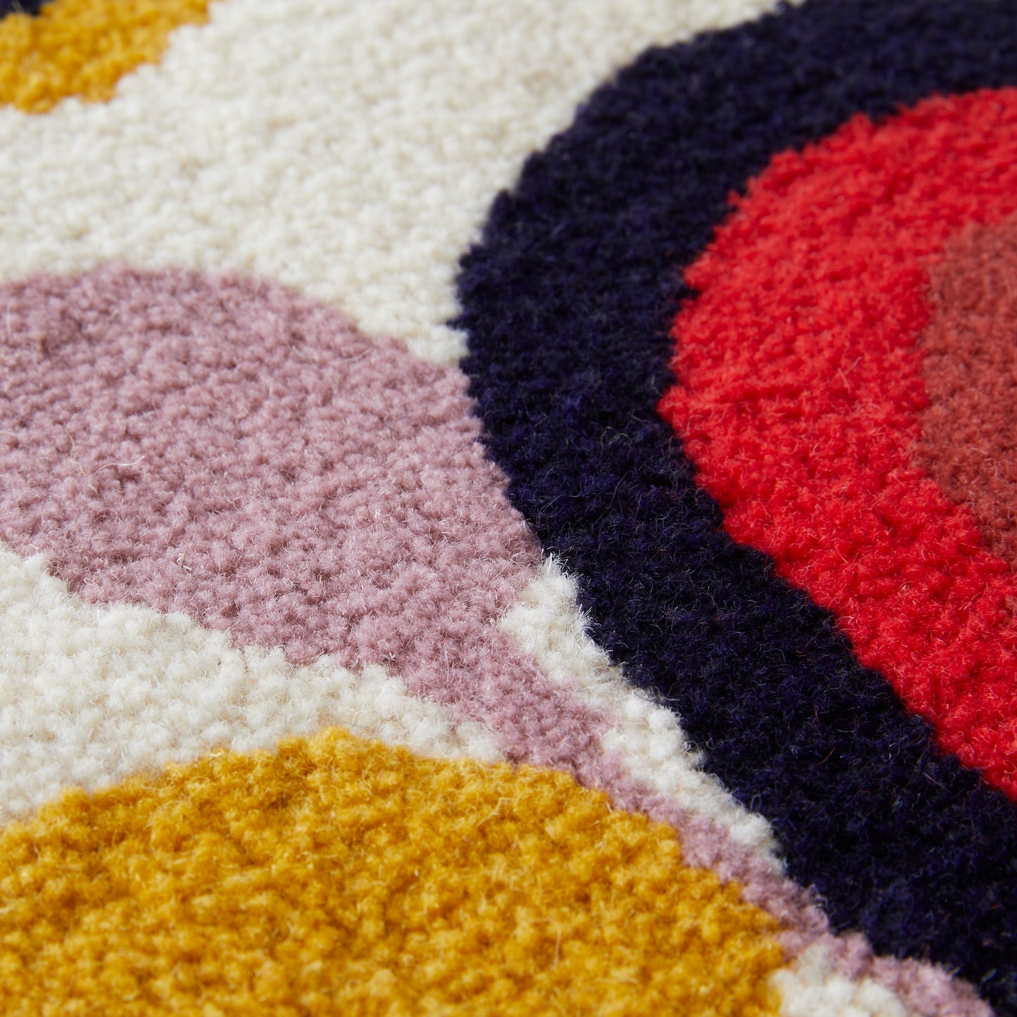 A cartoon-esque Candy Landscape rug runner with rainbows, hills and trees, hand tufted in a mixture of mustard, lilac, cream, red, coral, black, navy and maroon reclaimed yarns. A cut pile finish with hand bound mustard edges close up.