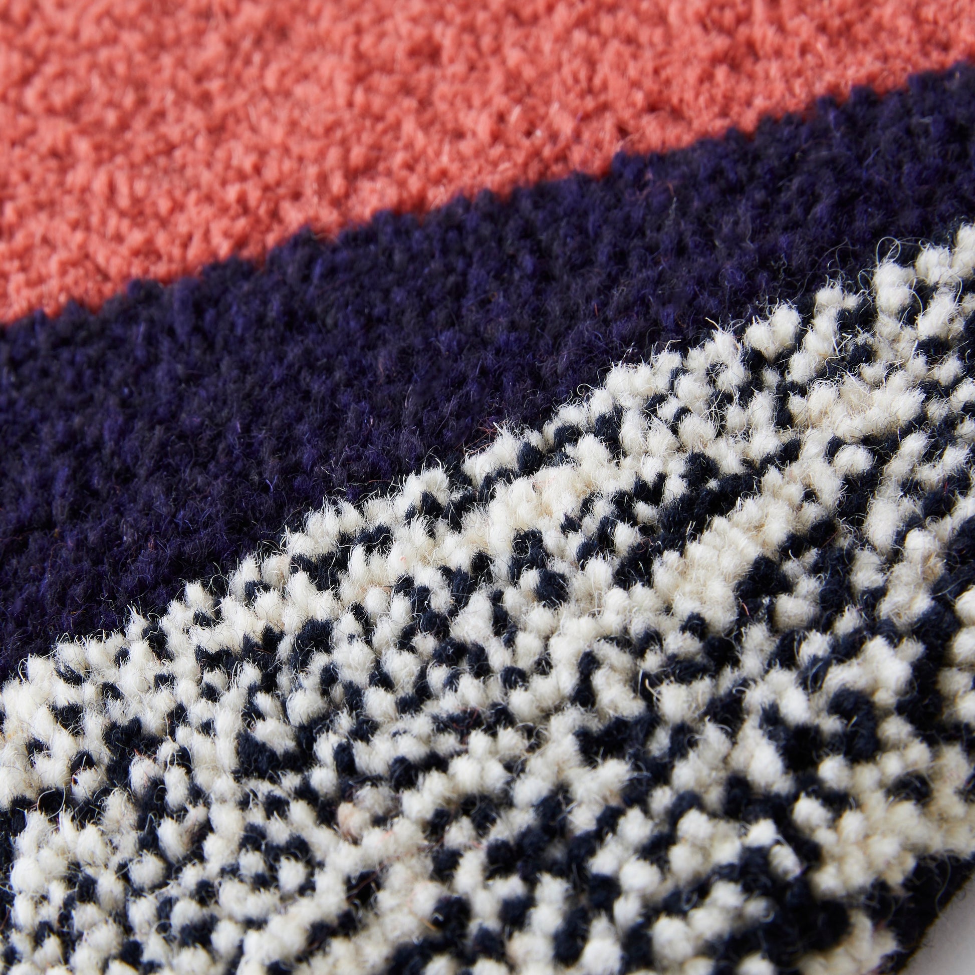 Handmade rug runner created using reclaimed yarns. Using Triangular fragments in Coral, Purple, Black and White flecks, Navy and Yellow with a hand bound dusky coral/pink edge close up.