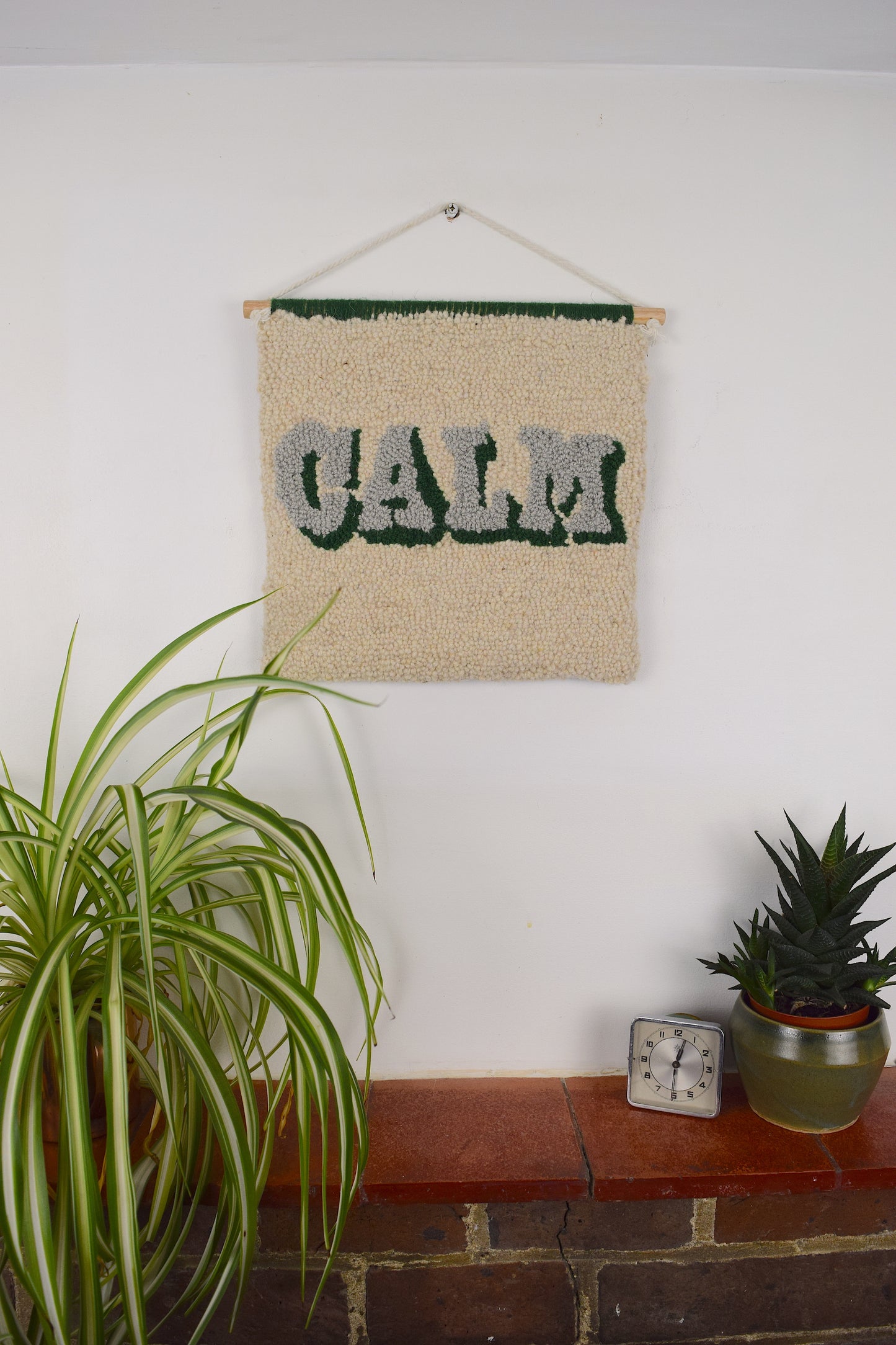 A vintage style typography Wall Hanging with the word CALM soft cream, khaki and green in a loop pile tuft using reclaimed yarn styled on a white wall with plants on the shelf below.